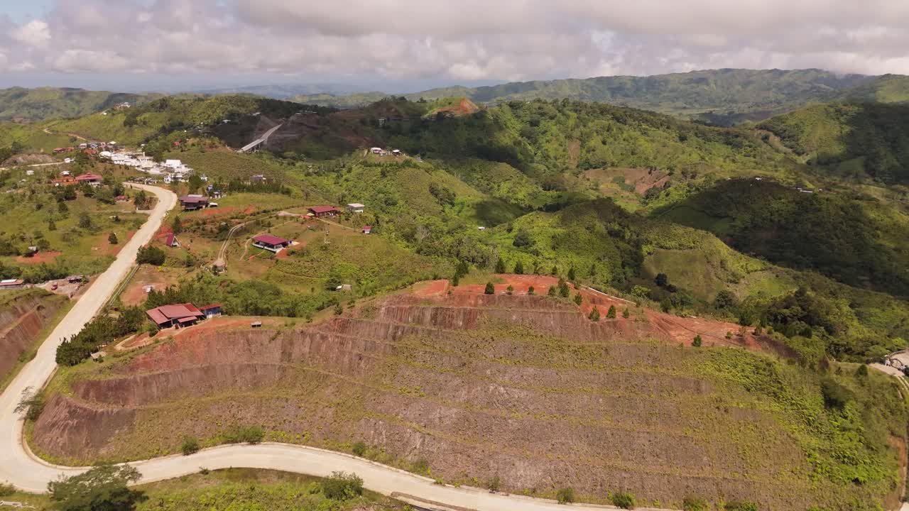 Drone footage of a winding mountain road passing through a rural hillside community in the Philippines, surrounded by layered terrain, lush greenery, scattered homes, bright daylight,rolling valleys