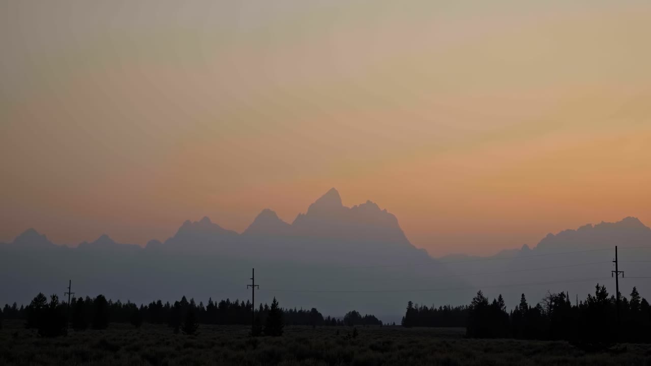 Stunning landscape nature tilting down shot of the Grant Teton mountain range during a hazy summer sunset with pine trees and power lines below near Jackson Hole, Wyoming, USA.