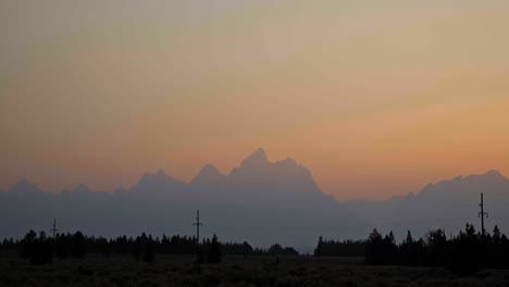 Stunning landscape nature tilting down shot of the Grant Teton mountain range during a hazy summer sunset with pine trees and power lines below near Jackson Hole, Wyoming, USA.
