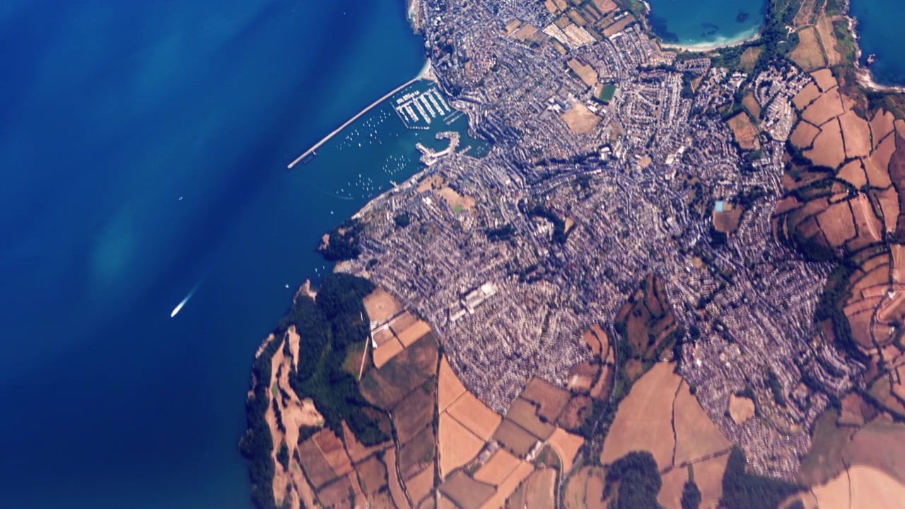 Città di Brixham nel Regno Unito vista dall'alto, che mostra il paesaggio costiero britannico con porto, case ed edifici e campi ingialliti circostanti.