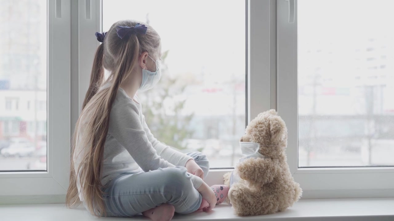 Junges Mädchen mit Gesichtsmaske sitzt am Fenster mit ihrem Teddybär