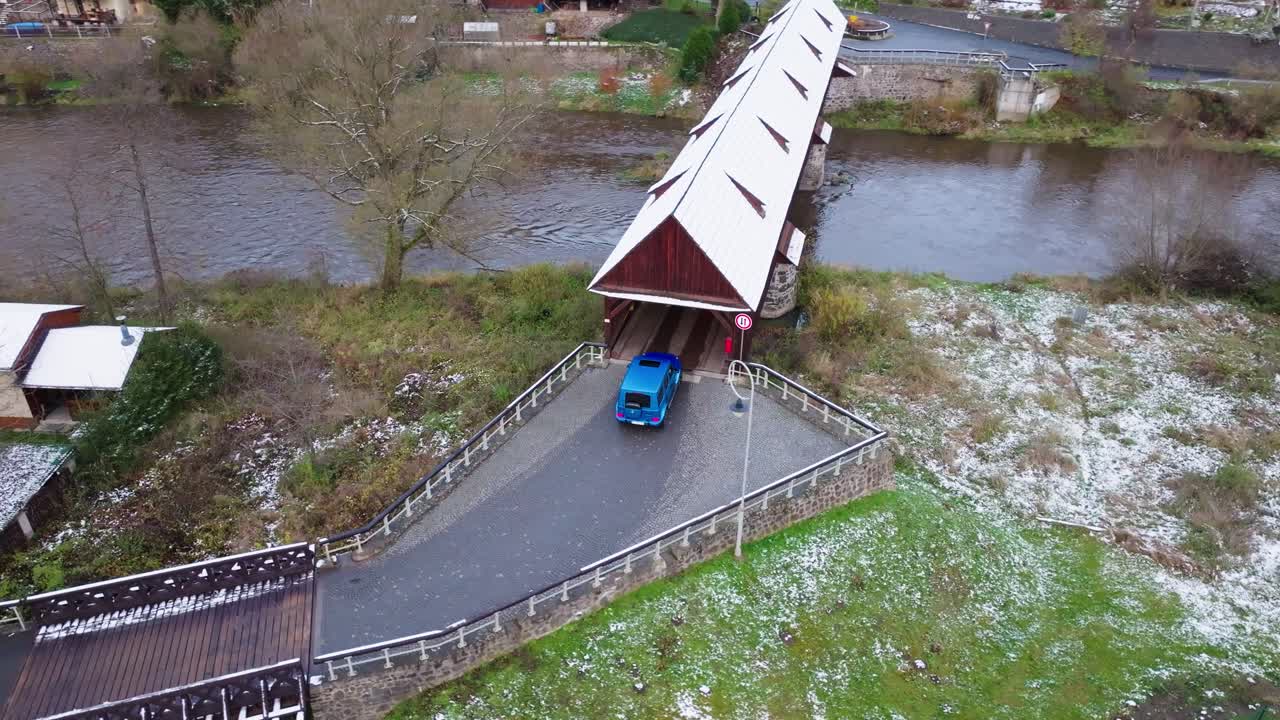 Aerial view of blue car entering covered bridge over river in winter, Czechia