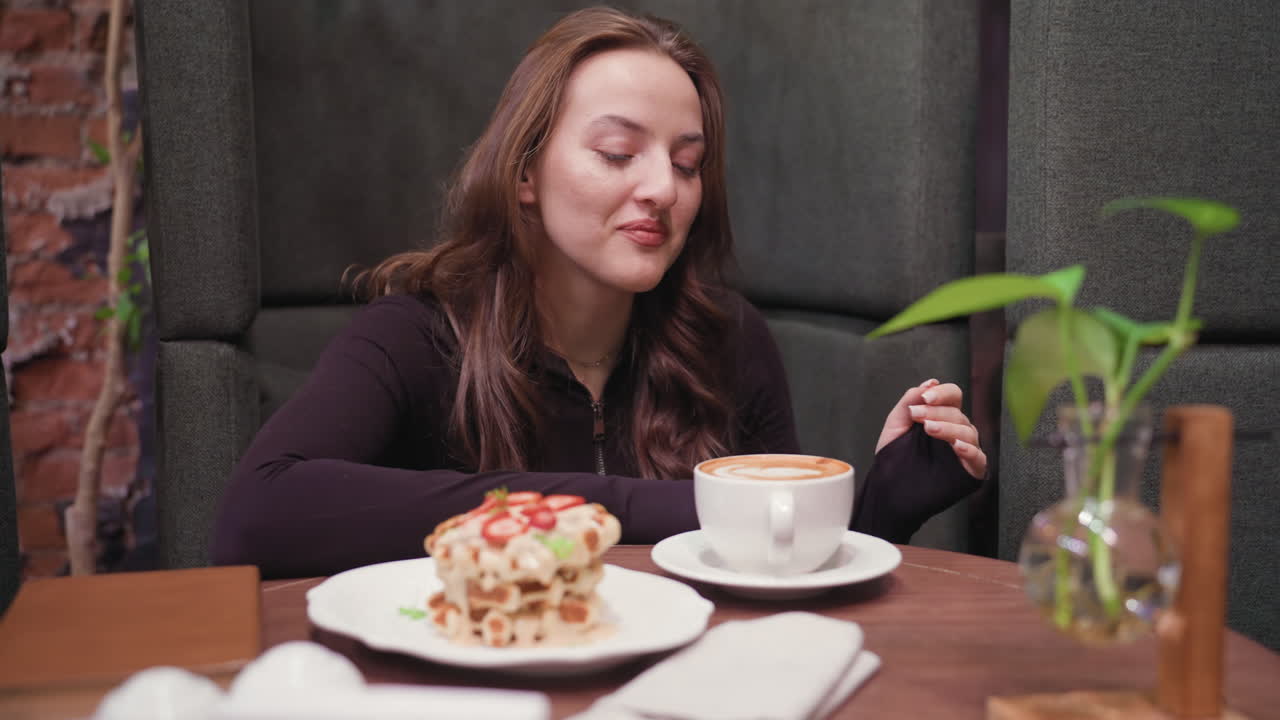 Young woman seated at wooden table gently leans forward with eyes closed, savoring aroma of latte in white cup, with waffles topped with strawberries beside her and plants in background