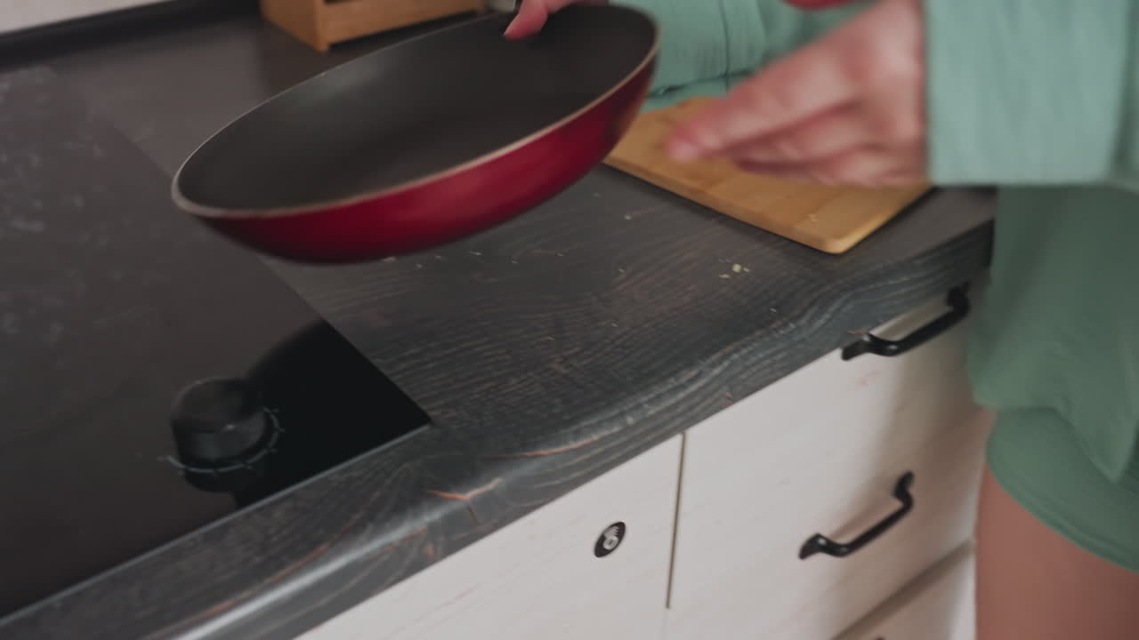 woman in green shirt opening lower kitchen drawer to take frying pan from storage, preparing to place it on electric stove with cutting board resting on dark countertop in modern indoor kitchen