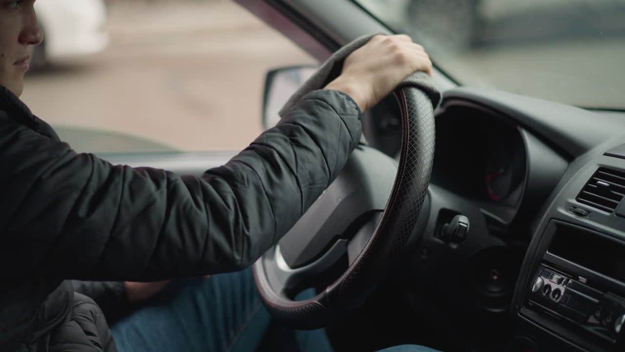 Side view of road traveler seated in car with window down using towel to wipe leather steering wheel in circular motion while vehicles pass by on street through blurred background scene