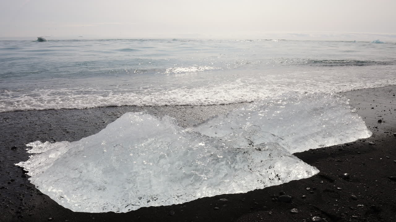 Ocean waves lapping up chunks of ice sparkling on a black sand beach, handheld