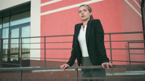 jonge kampioen in zwarte blazer rust handen op ijzeren reling buiten residentieel gebouw geverfd rood, straalt vertrouwen en kalmte met focus op moderne architectuur en stedelijke omgeving in zachte verlichting