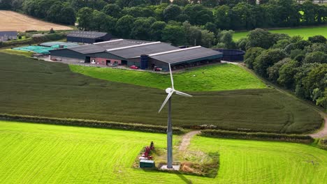 aerial footage shows a wind turbine spinning in a sunlit rural field near farm buildings in kinross, scotland. bright daylight, smooth camera movement