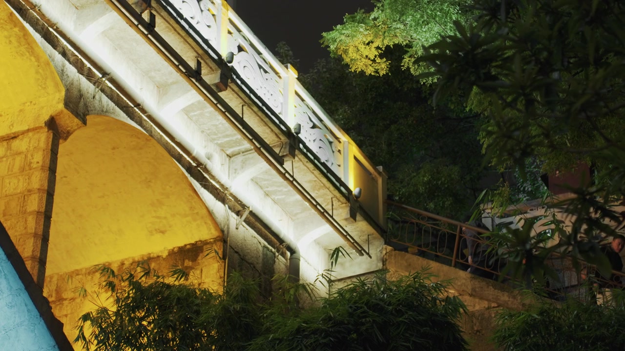 Escalera iluminada para caminar de noche, luces blancas frías trazan un camino ascendente a través de árboles sombreados, escalones de hormigón y barandilla de metal, sotobosque y bordes de jardín, eco de pasos, ambiente urbano misterioso.
