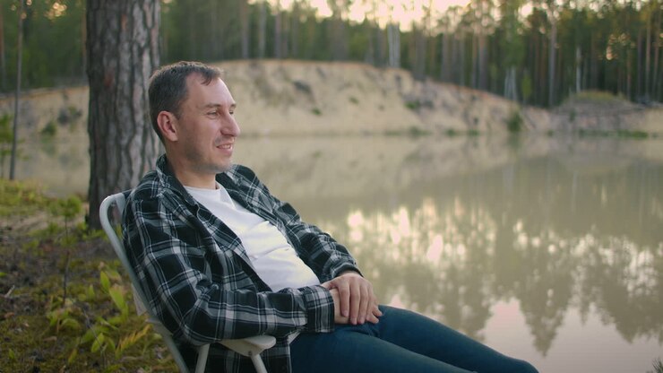uomo adulto rilassato è seduto in poltrona sulla riva di un calmo lago pittoresco nella foresta e ammirando la natura e la calma