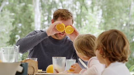 Padre-Haciendo-Reír-A-Los-Niños-En-La-Mesa-Del-Desayuno-Haciendo-Caras-Con-Naranjas-Cortadas-Para-Los-Ojos