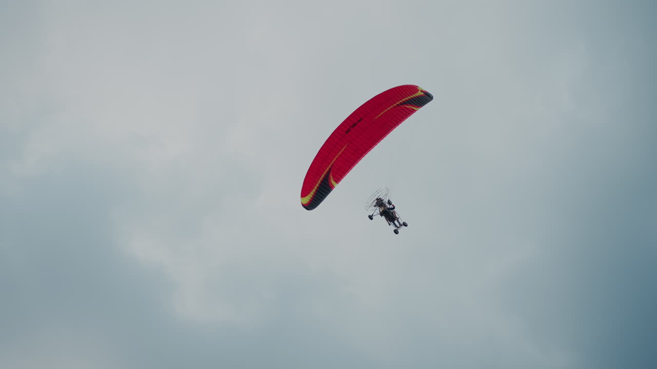 low angle capture of powered paraglider soaring overhead with red canopy against cloudy sky, pilot visible under wing in serene aerial scene emphasizing height, motion