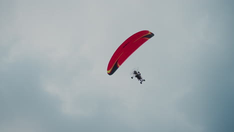 low angle capture of powered paraglider soaring overhead with red canopy against cloudy sky, pilot visible under wing in serene aerial scene emphasizing height, motion