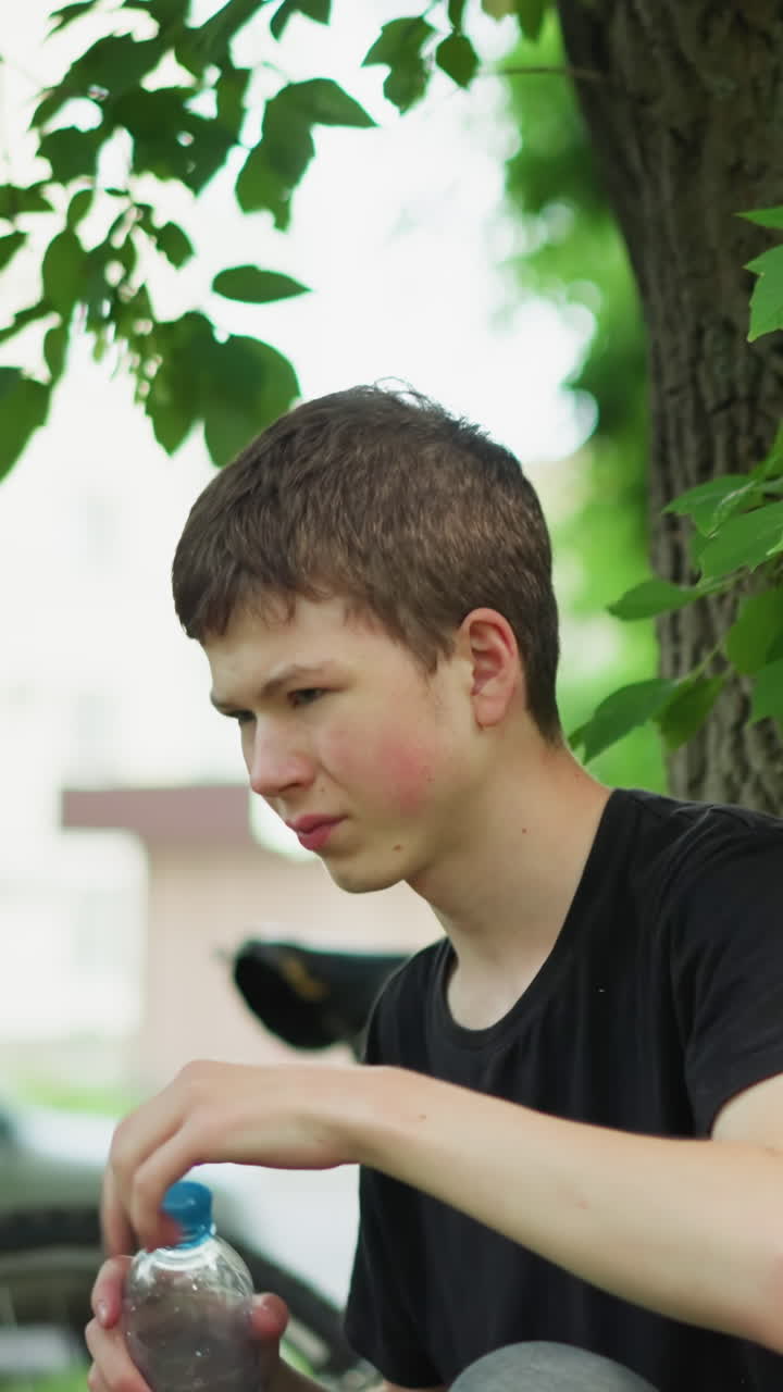 어린 소년은 자전거 타고 나서 나무 에서 휴식을 취하는 동안 물을 마시며, 자전거는 울창한 초록색으로 둘러싸여 근처에 주차되어 있습니다. 배경에서 지나가는 차량의 흐릿한 시각