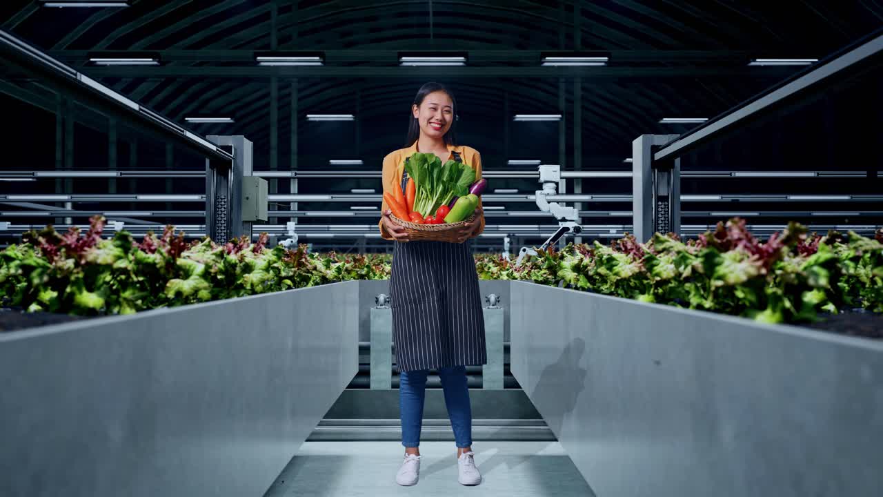 cuerpo lleno de agricultora asiática llevando una canasta de verduras sonriendo cálidamente en la granja inteligente