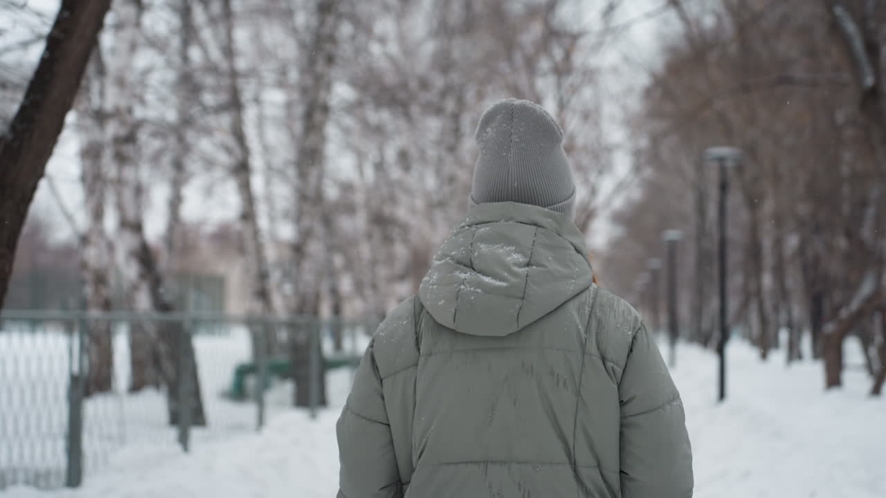 Dog owner standing on snowy path during winter, wearing gray beanie and puffy jacket, seen from behind, snowflakes visible on clothing, surrounded by leafless trees and soft light in calm cold outdoor setting