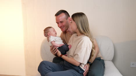 beautiful caucasian couple sitting on the sofa with their newborn. happy dad kisses the sleeping infant son.