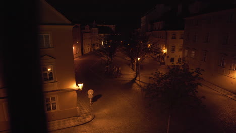 Small-empty-city-square-Kampa-in-Prague,-Czechia,-with-trees-and-deserted-streets-during-a-Covid-19-lockdown-at-night,-houses-and-pavement-lit-by-golden-street-lantern-light,shot-through-iron-railing