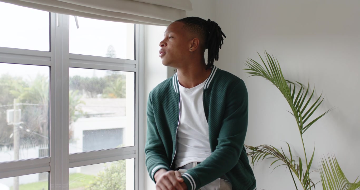 Afroamerikanischer Mann sitzt auf der Fensterbank, bedeckt sein Gesicht, senkt die Hand, dem Fenster zugewandt, nachdenklich