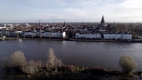 Steady-aerial-view-of-white-countenance-facade-of-tower-town-Zutphen-rising-behind-in-The-Netherlands-with-river-IJssel-strong-current-passing-by-the-Dutch-city