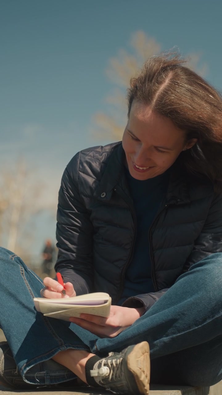 Primo piano di una donna seduta all'aperto in una giornata di sole, che tiene in mano un quaderno aperto e una penna rossa, con un'espressione pensierosa e il vento che le scompiglia i capelli. La donna indossa abiti casual, inclusi jeans e una giacca.