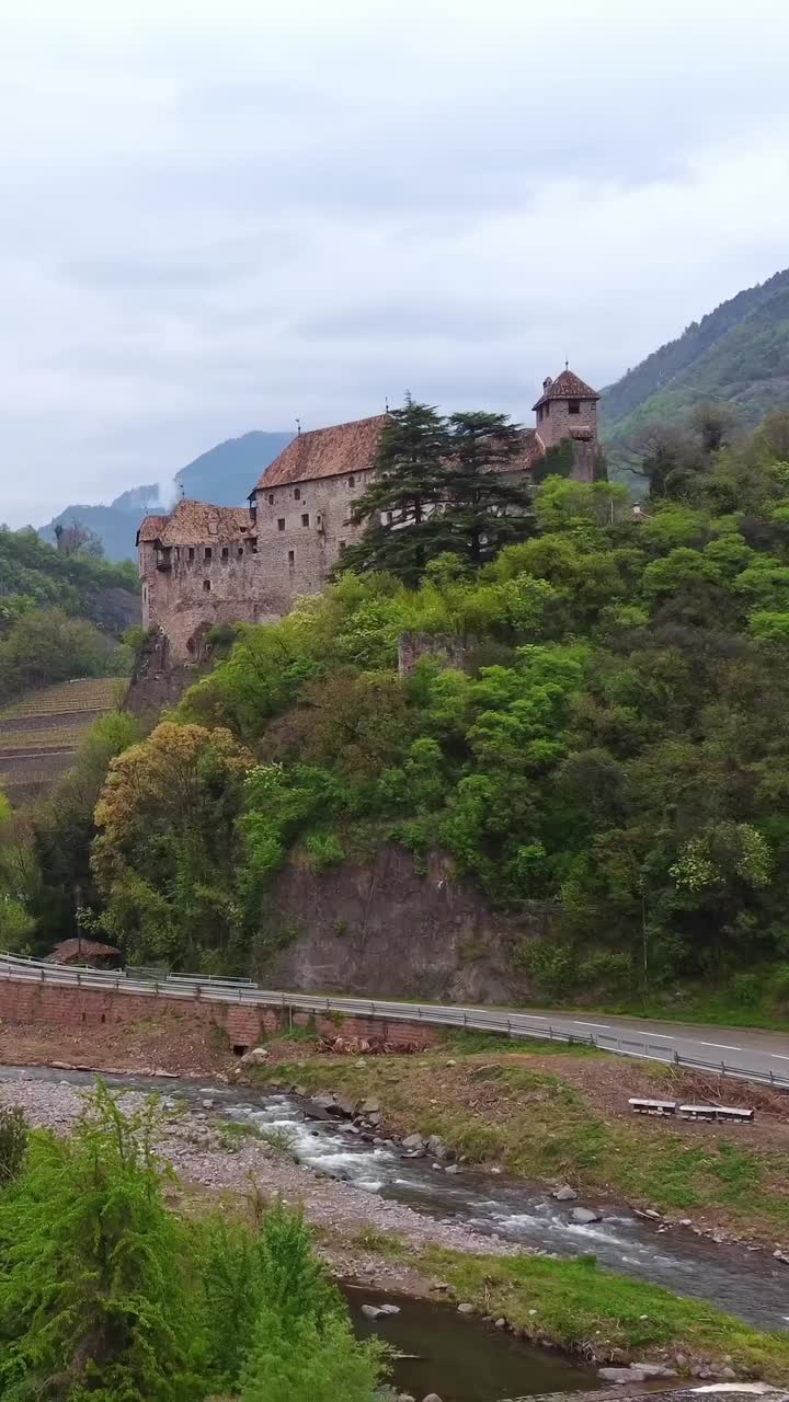 Vertical drone flight near Roncolo Castle showing a street and small river in front, framed by nature