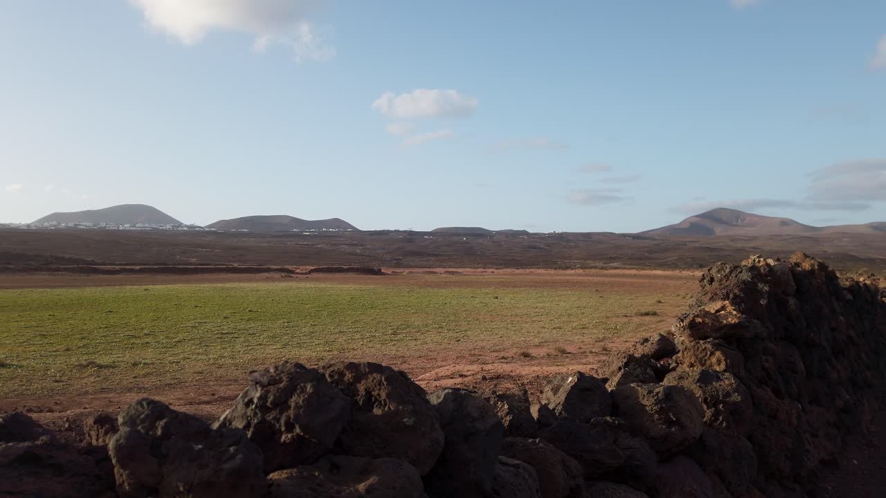 Volcanic rocks border a green meadow with mountains and a small village in the distance, under a blue sky with some clouds, in lanzarote, canary islands