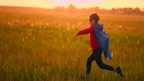 un niño con un traje y una máscara de superhéroe corre a través del campo al atardecer en la hierba soñando e imaginándose a sí mismo un héroe y un ganador. familia divertida y feliz