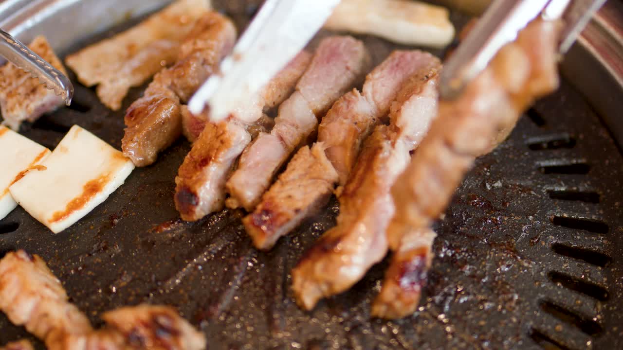Hand uses metal tongs to turn sizzling pork belly slices on hot Korean barbecue grill
