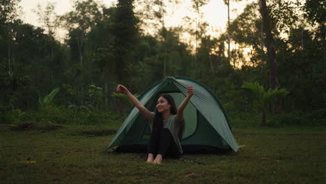 young woman relaxing by her tent in a forest at sunset