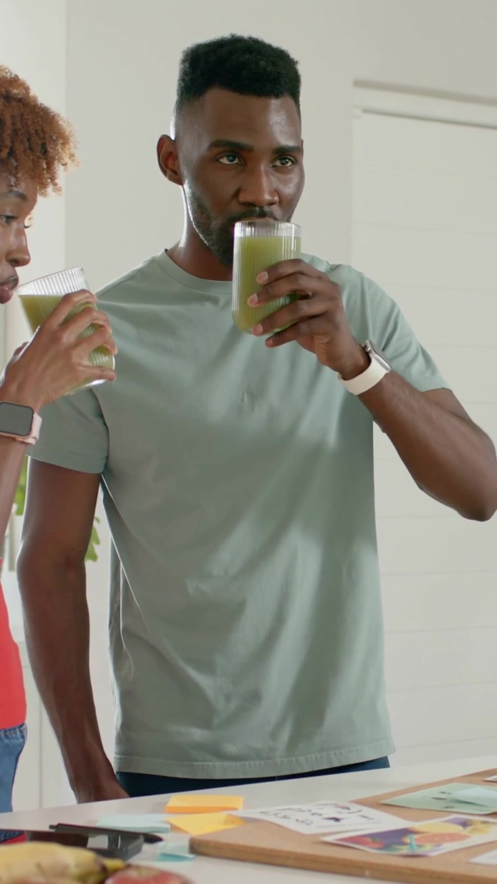 Vidéo verticale : Homme en t-shirt vert d'eau dégustant un smoothie vert au comptoir de la cuisine, femme levant un verre.