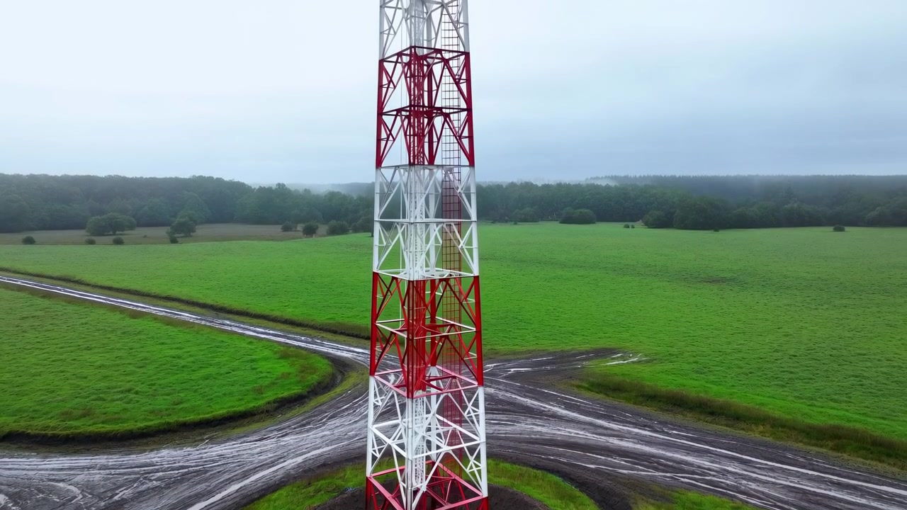 Pemandangan udara menara kisi baja tinggi merah dan putih yang berdiri di padang rumput hijau, dengan jalur akses melingkar berlumpur, tanah basah, genangan air, dan cahaya mendung yang menyebar di sekitar lokasi pedesaan, dengan menara tersebut