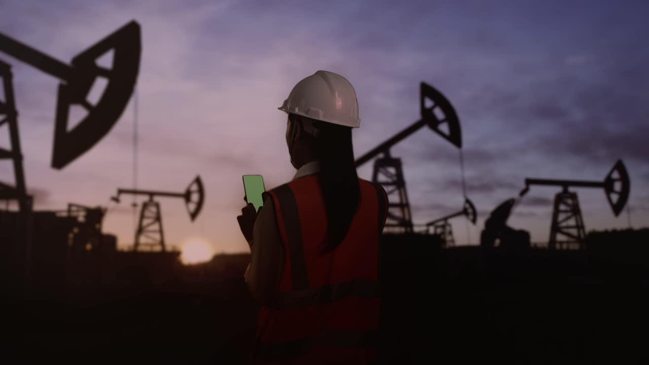 vista trasera de una ingeniera asiática con casco de seguridad usando un teléfono inteligente de pantalla verde inspecciona las bombas de petróleo al amanecer en un gran campo petrolífero