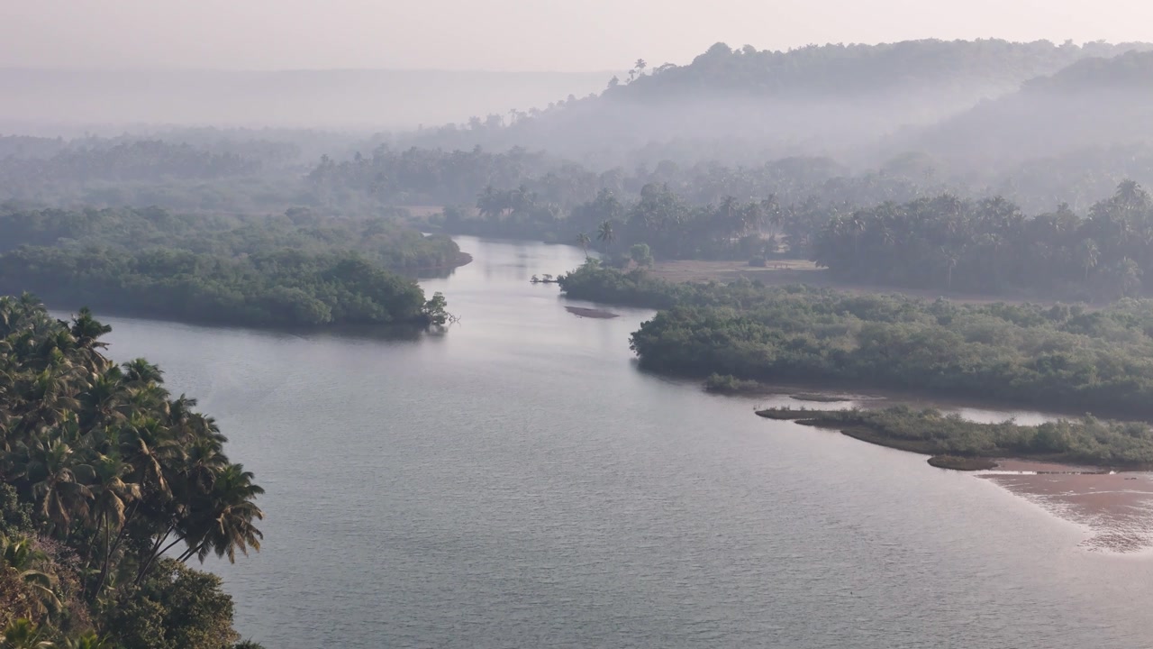 Misty morning aerial view of a wide river winding through a dense tropical jungle valley in the Konkan region of India.