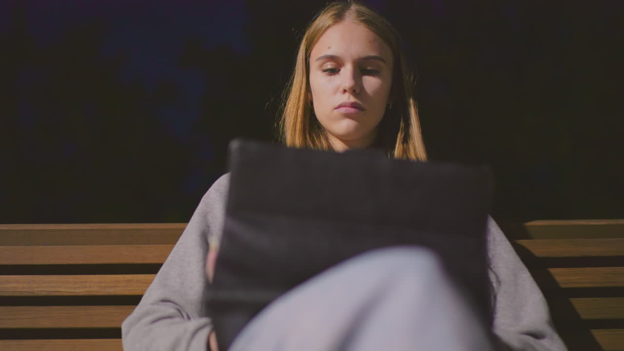 Close-up of girl sitting on park bench at night, fully focused on tablet under soft park lighting, with a serene deep blue sky in background