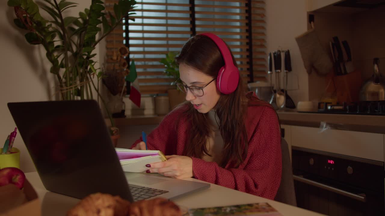 A confident brunette girl in red wireless headphones and a sweater studies foreign languages taking notes from a lesson in her notebook while sitting in the kitchen in a modern apartment near the flag of Italy