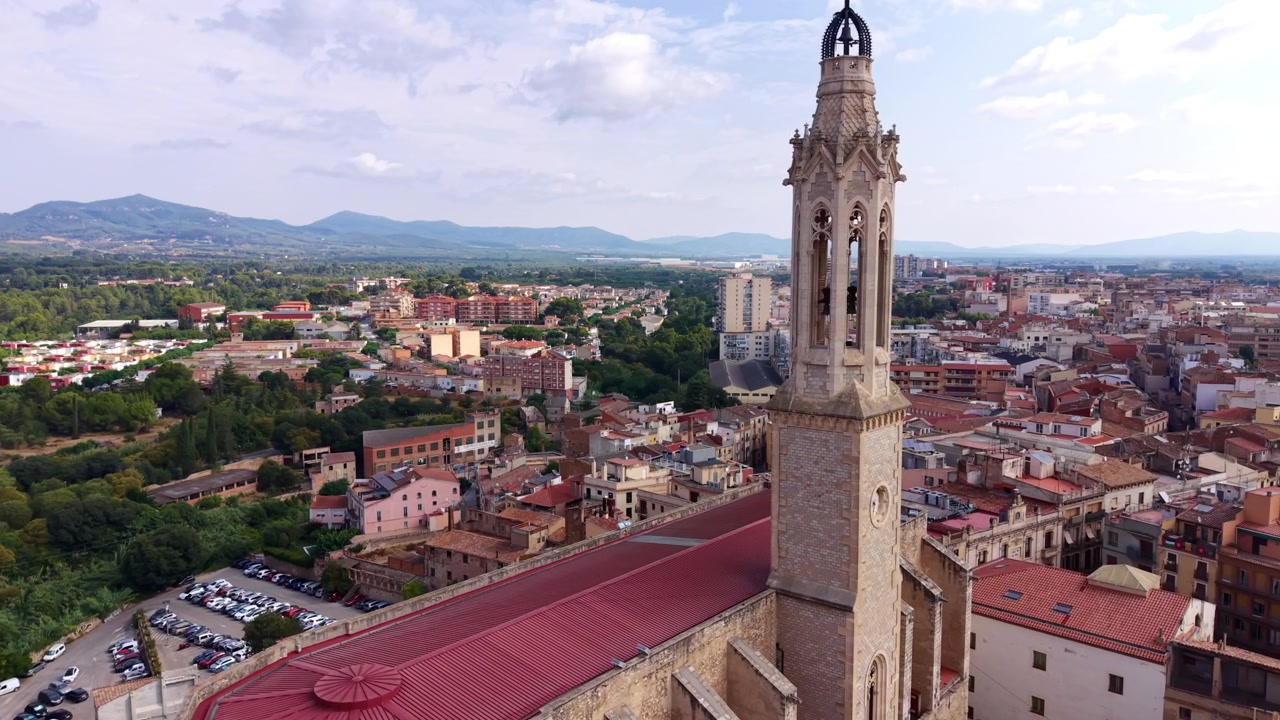 Un campanario de iglesia gótica se eleva sobre un pueblo español con tejados de terracota, edificios residenciales, exuberantes árboles verdes y una cordillera que se extiende hasta el horizonte.