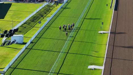 multiple jockeys ride horses along a green turf racetrack, captured from above in bright daylight with steady drone movement and clear shadows