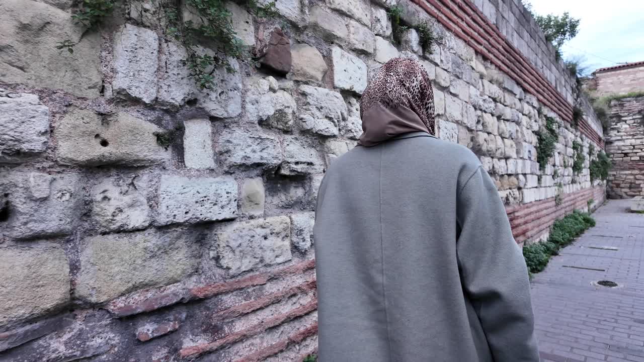 A woman in a hijab walks down a street lined with stone walls