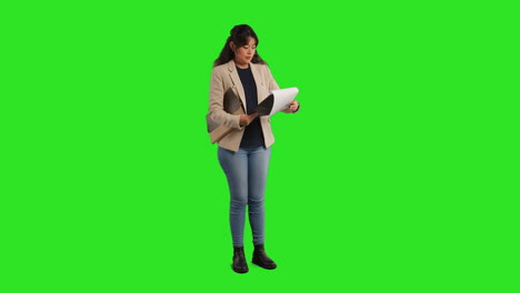 Full-Length-Studio-Portrait-Of-Female-Teacher-Or-Businesswoman-Standing-Against-Green-Screen-Looking-At-Clipboard