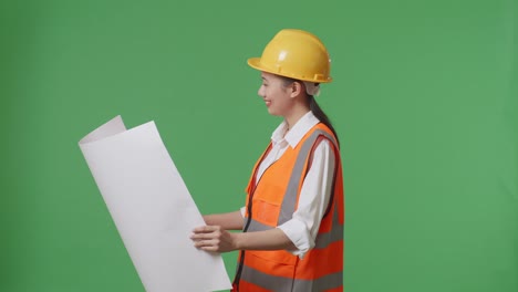 side view of asian female engineer with safety helmet looking at blueprint in her hands and looking around while standing in the green screen background studio