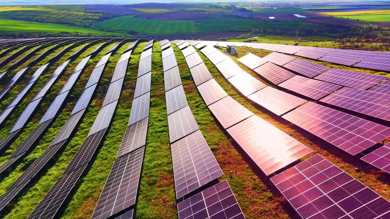 Luftfoto af buede rækker af solpaneler. Nærbillede fra høj vinkel af lange fotovoltaiske anlæg på en grøn bjergskråning med stærk solrefleksion og fjerntliggende landbrugsjord.