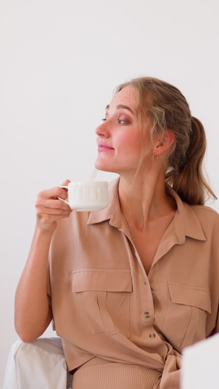 Young woman in beige shirt enjoys cup of hot drink and smiles putting hand on closed laptop at office table after working day slow motion