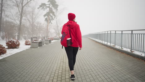 vista trasera de una mujer con chaqueta roja de invierno y botas, llevando una mochila con auriculares en el cuello, caminando por un camino cubierto de nieve rodeado de bancos y árboles en una atmósfera de invierno brumosa y serena