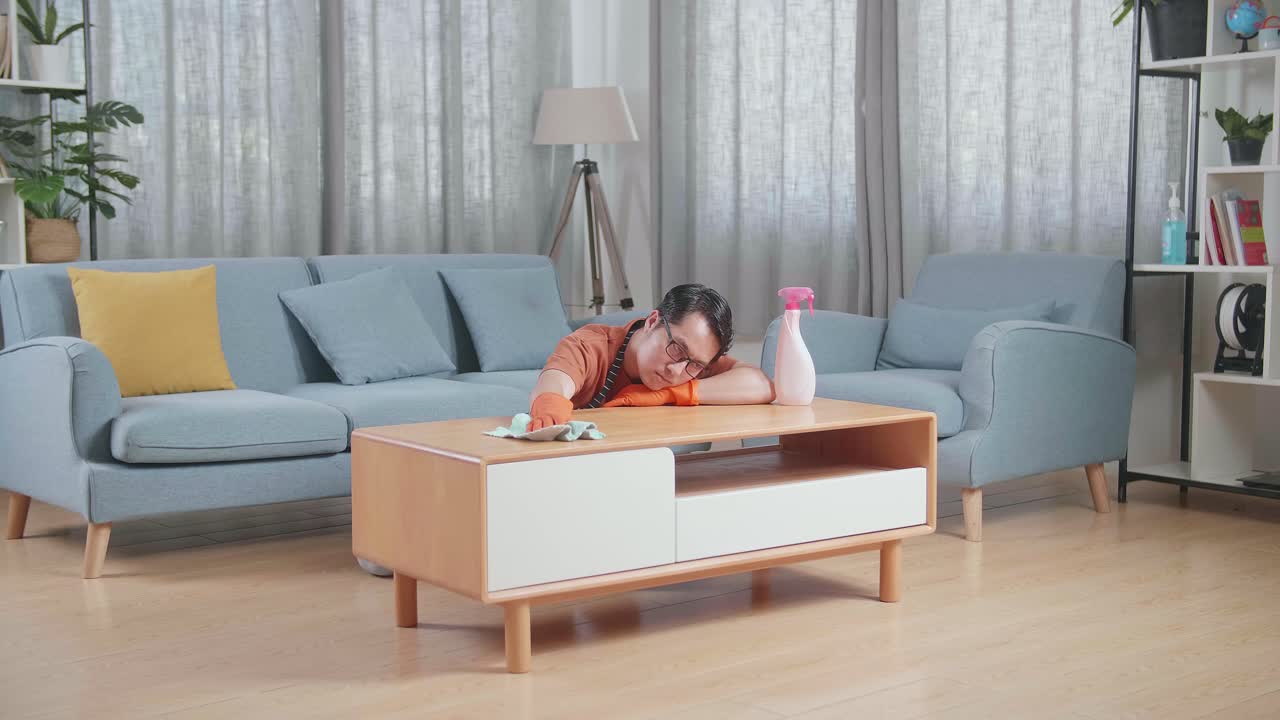 지친 아시아 남성 집주인이 집에서 스프레이로 테이블을 청소하고 있습니다.
