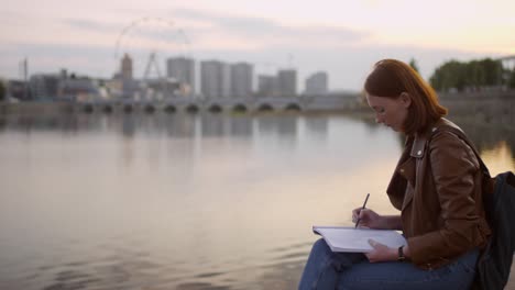 Pretty-Ginger-Young-Woman-With-Freckles-Sitting-On-Embankment-And-Drawing-Cityscape