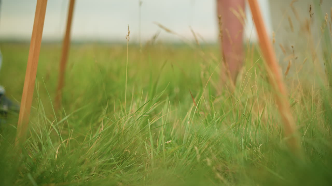 A close-up shot of the legs of a wooden tripod standing in a grassy field. The soft focus and natural setting evoke a sense of calm and simplicity in this peaceful scene