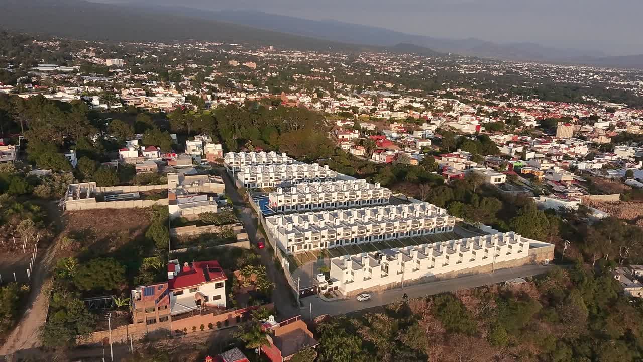 Aerial view of a newly built residential complex showcasing modern architecture. Nestled within a vast cityscape extending towards a mountainous backdrop bathed in the warm glow of late afternoon sun