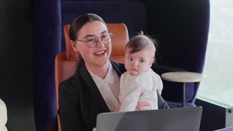 Close-up-of-a-confident-brunette-businesswoman-girl-in-round-glasses-and-in-business-clothes-sitting-at-the-office-desk-and-holding-her-small-baby-child-in-her-arms-during-a-business-meeting-in-the-office