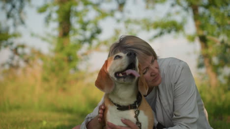 woman seated on grassy field holding her dog affectionately, gently patting and resting head on dog, creating touching moment of warmth and love, background features lush greenery and trees